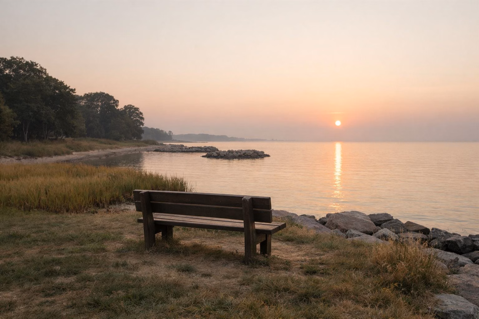 A quiet grassy point of land by calm open water at sunrise, a single wooden bench facing the horizon, soft haze