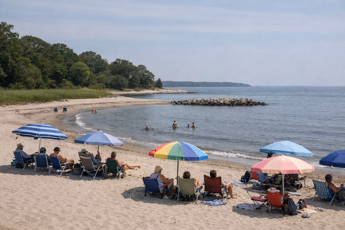 Beachgoers with umbrellas and chairs spread out on a wide sandy beach, small calm waves, low rock jetty in the distance