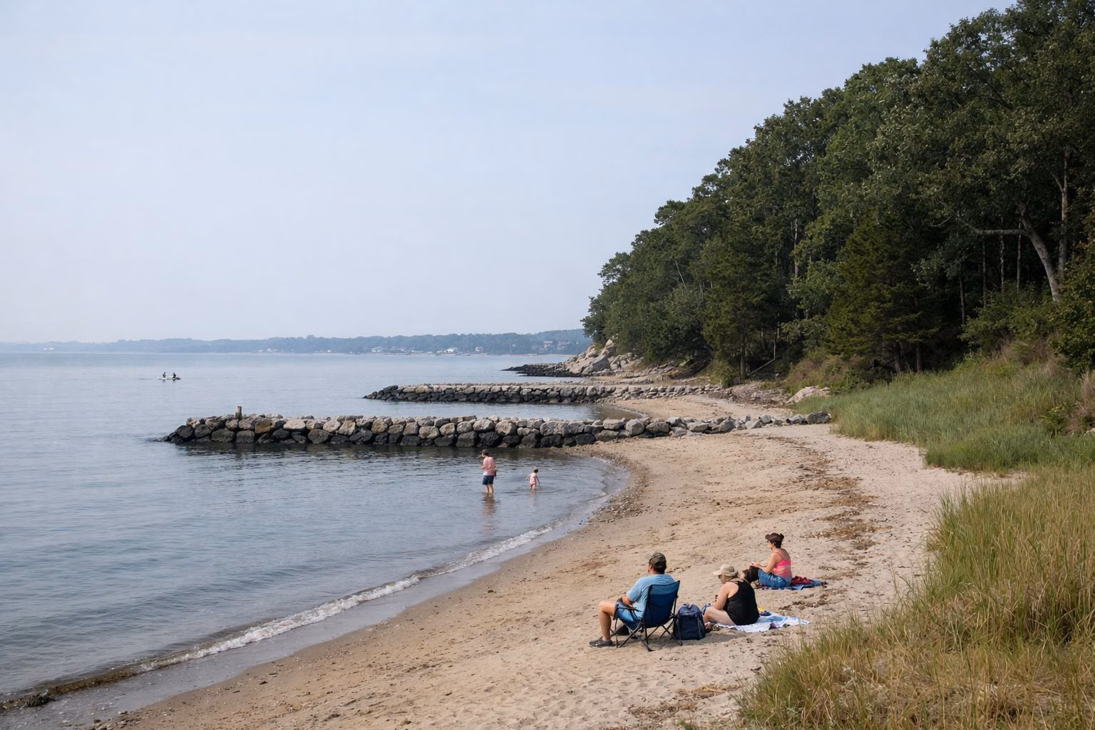 A small quiet sandy beach below a low wooded bluff, a few people, calm water