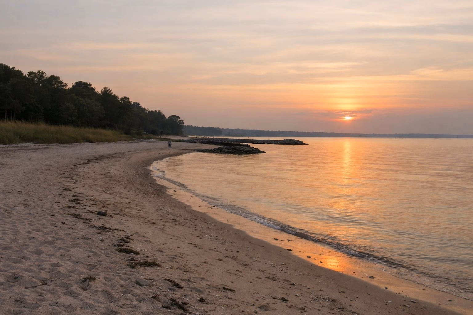 An empty sandy beach at sunset, calm water reflecting orange light, a lone walker far off