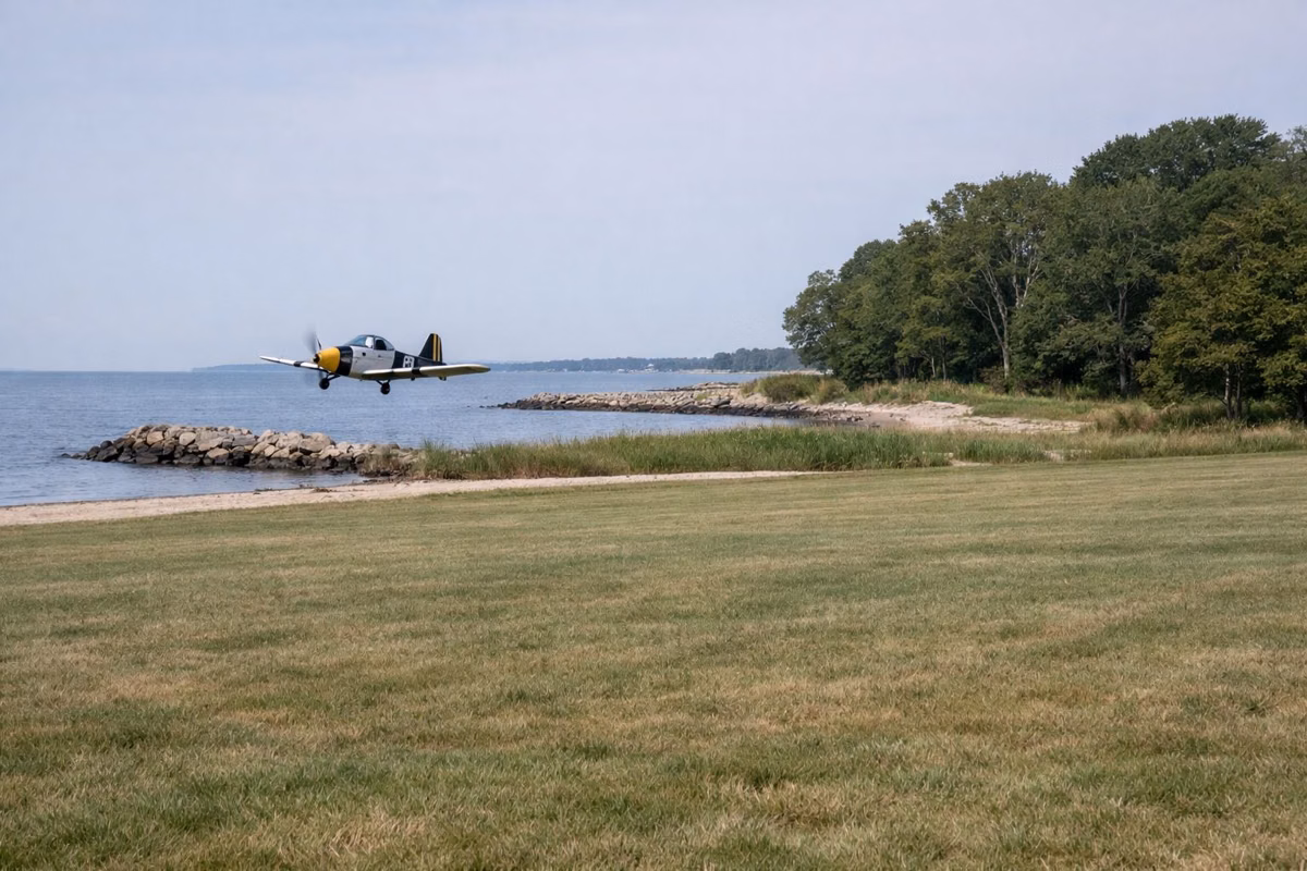 A small radio-controlled model airplane flying low over a wide mown grass field by the shore