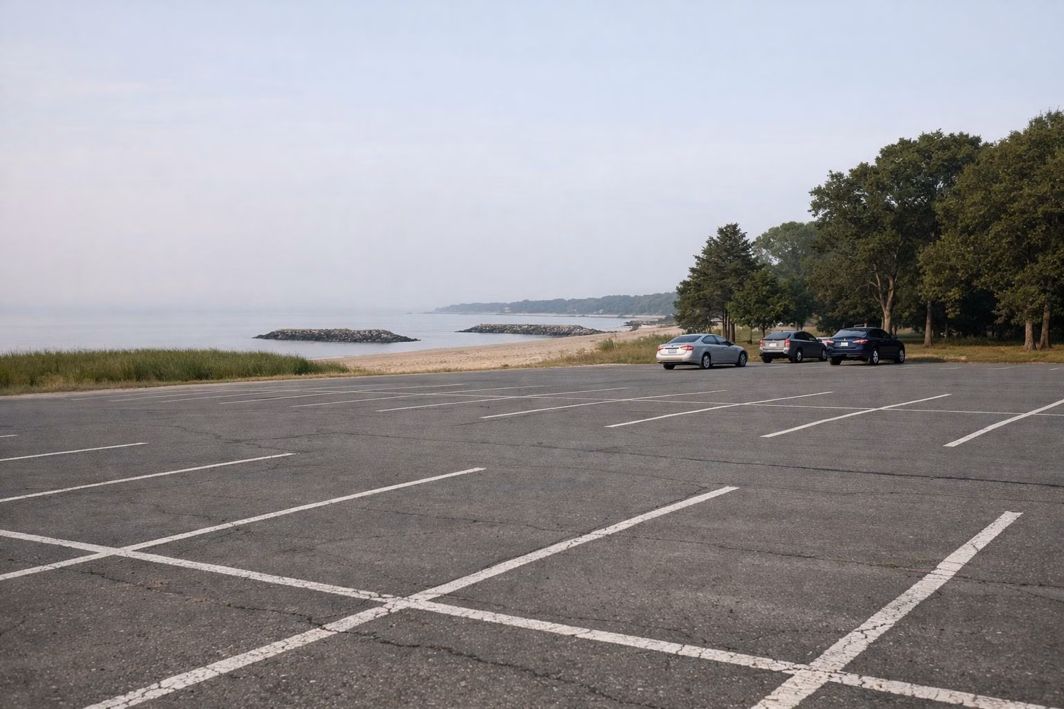 A wide paved beach parking lot in early morning with a few cars far off near the trees and the beach