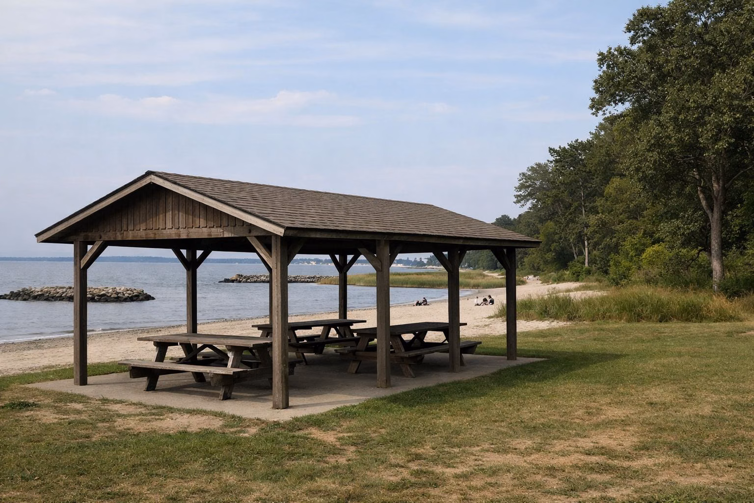 A simple open-sided wooden picnic shelter with picnic tables on a lawn near a sandy beach