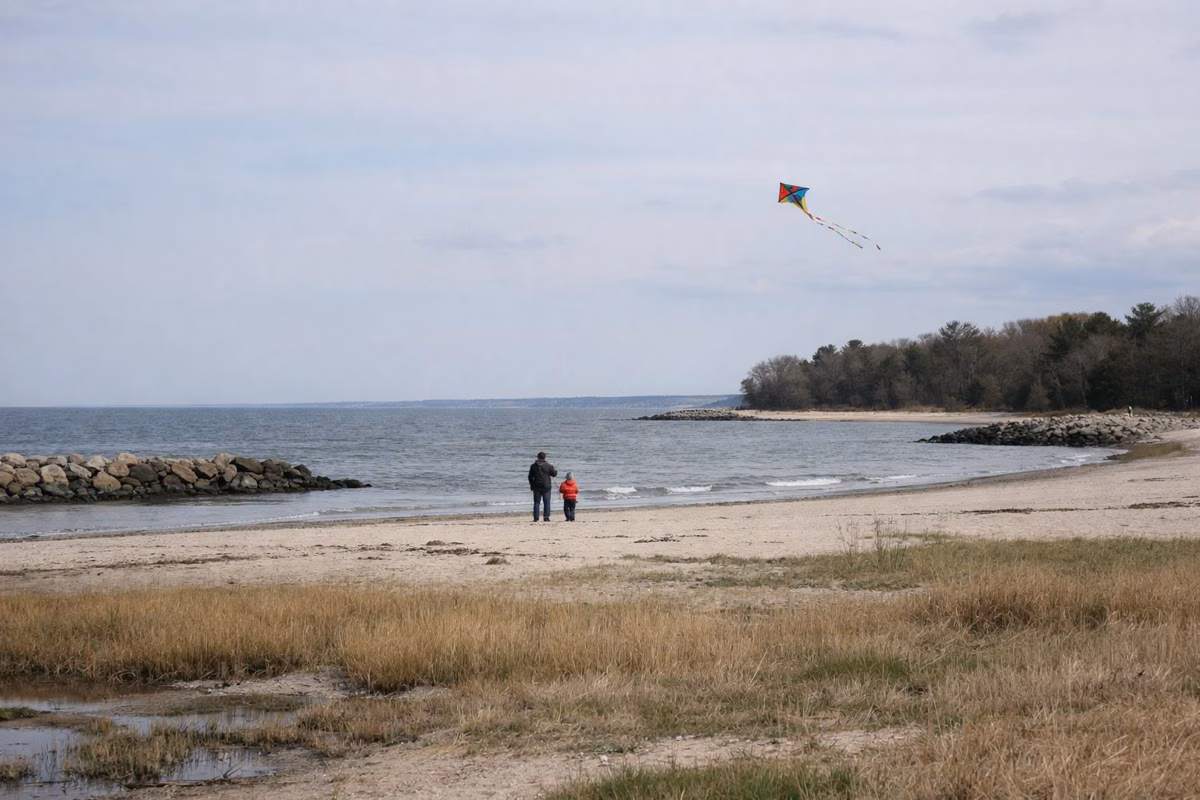 A parent and child flying a kite on a wide beach on a breezy day, seen from a distance