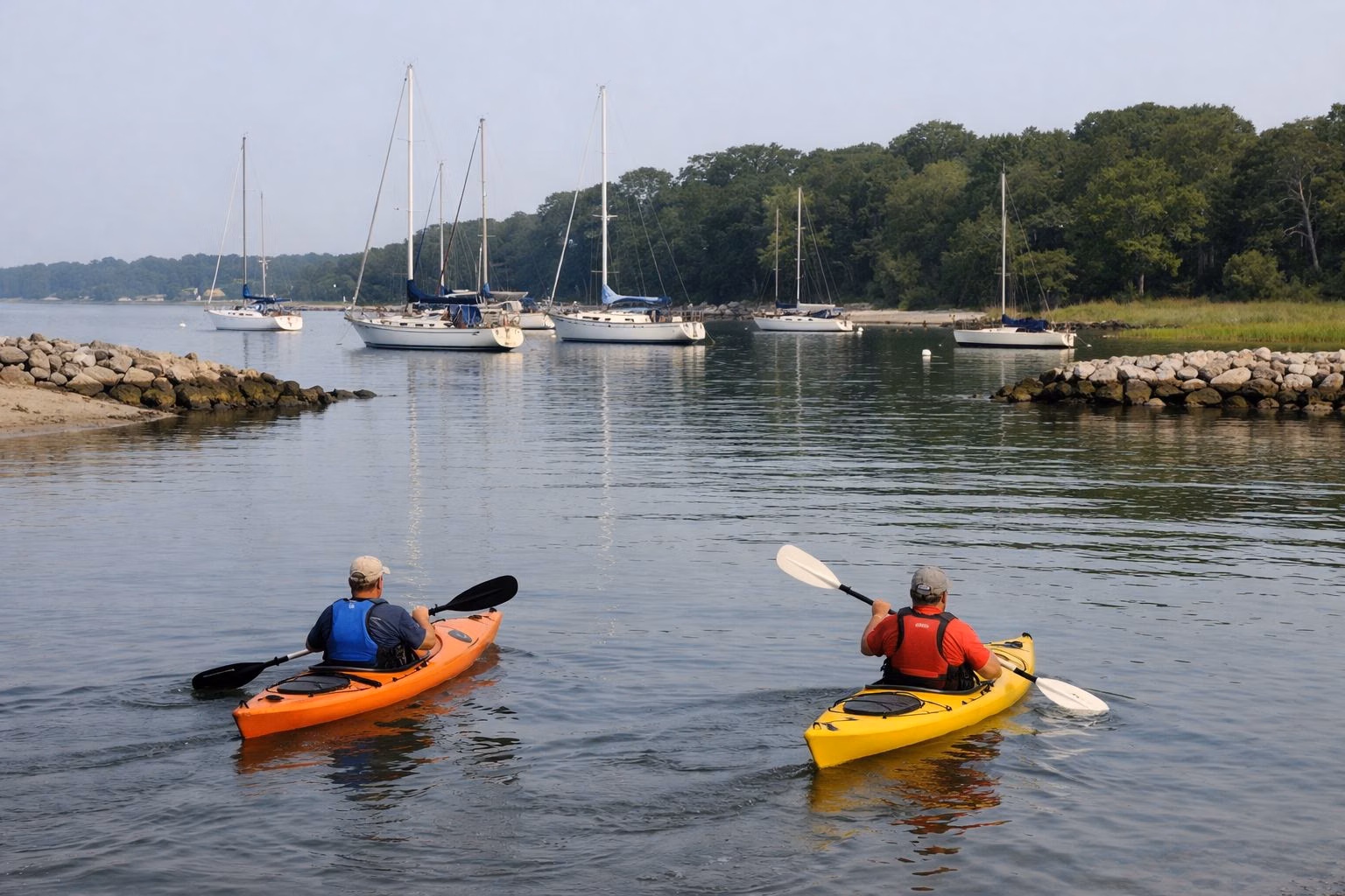 Two kayakers paddling on a calm tidal river mouth with moored sailboats behind